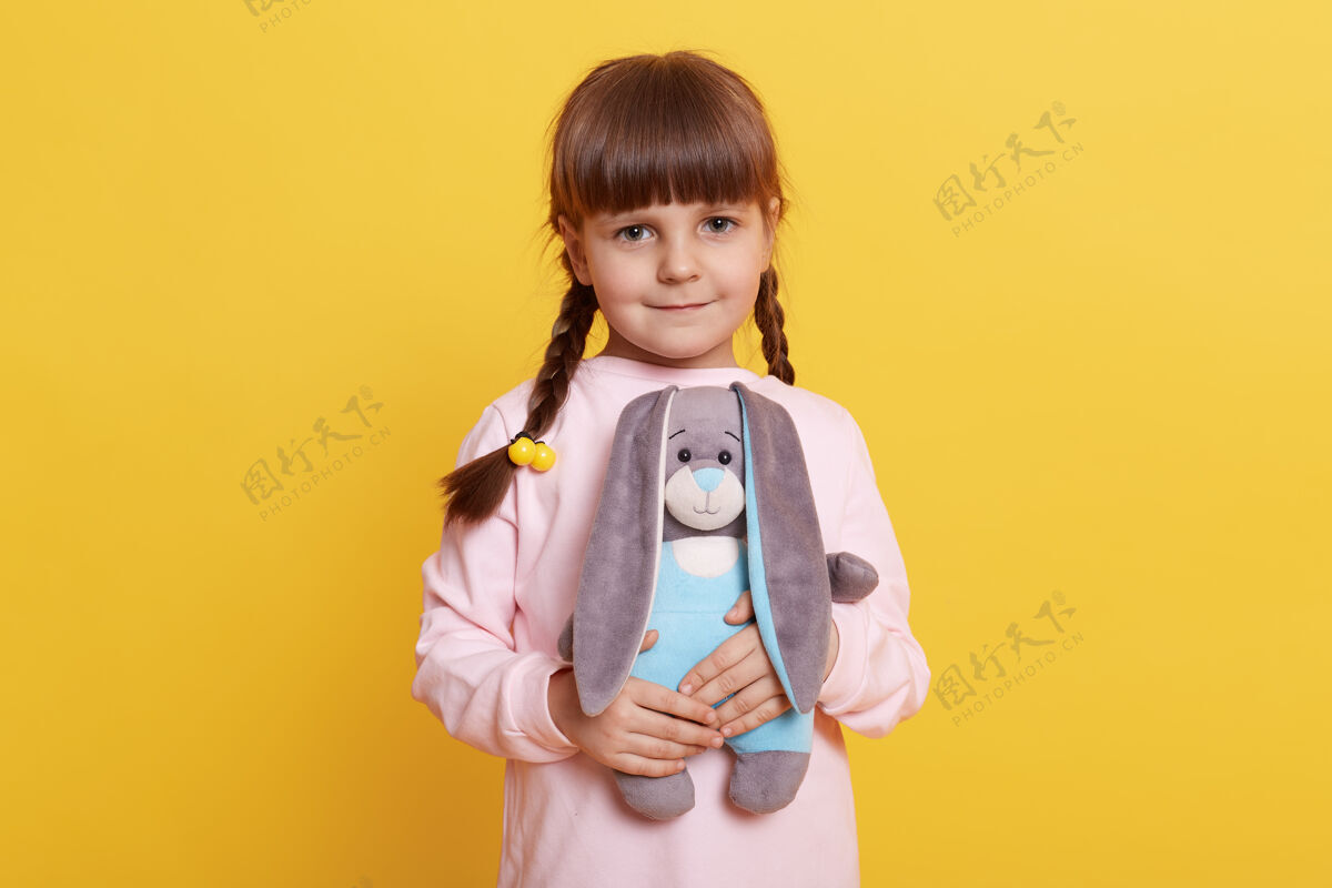 玩耍带着辫子微笑的女孩儿穿着浅猪衬衫 在黄色背景下摆出孤立的姿势 手里拿着柔软的兔子玩具 直视镜头 孩子在玩她的玩具温柔孩子欢呼