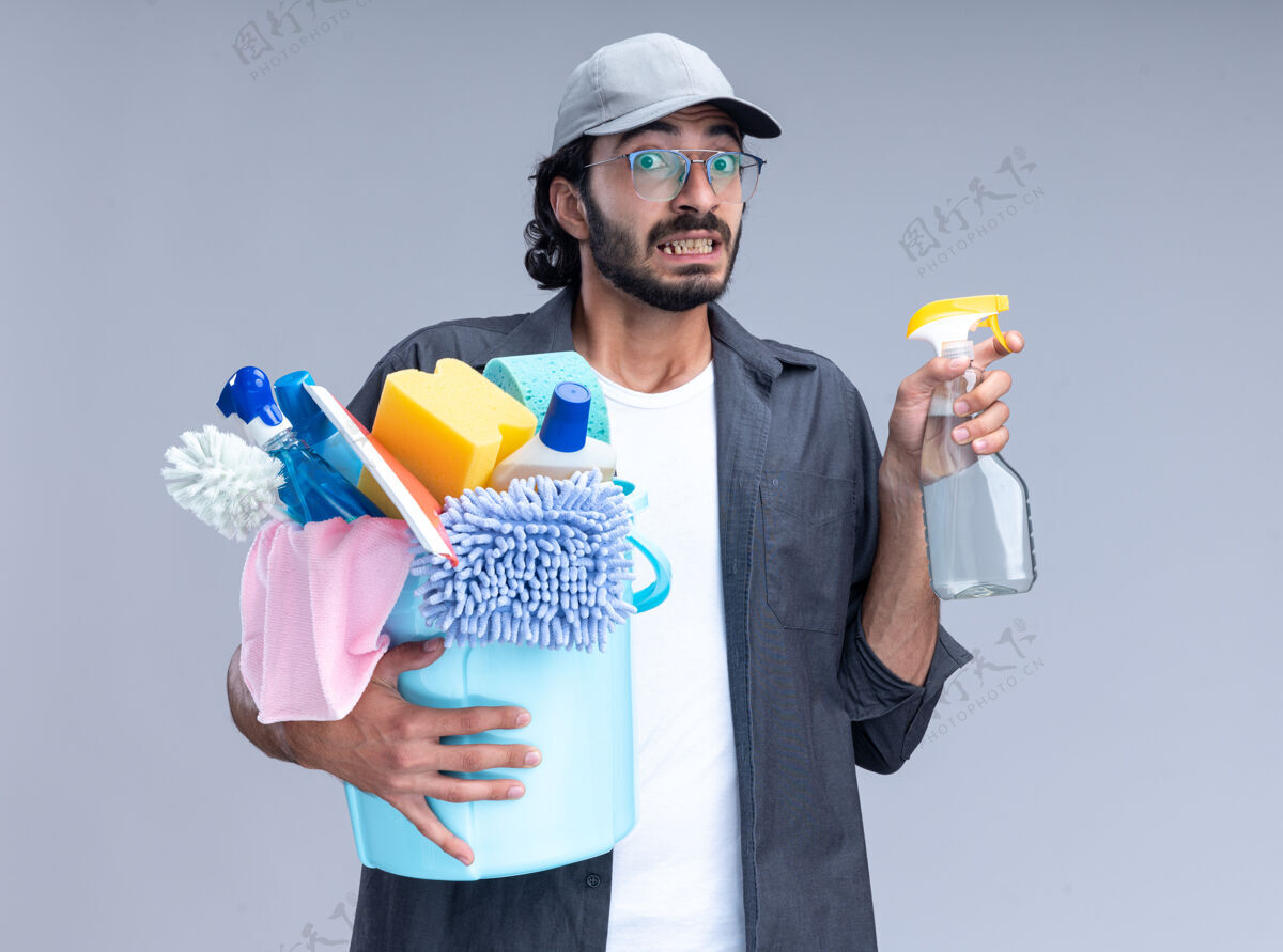 站着关心年轻帅气的保洁员 穿着t恤 戴着帽子 拿着一桶清洁工具 把喷雾瓶隔离在白色的墙上年轻瓶子清洁
