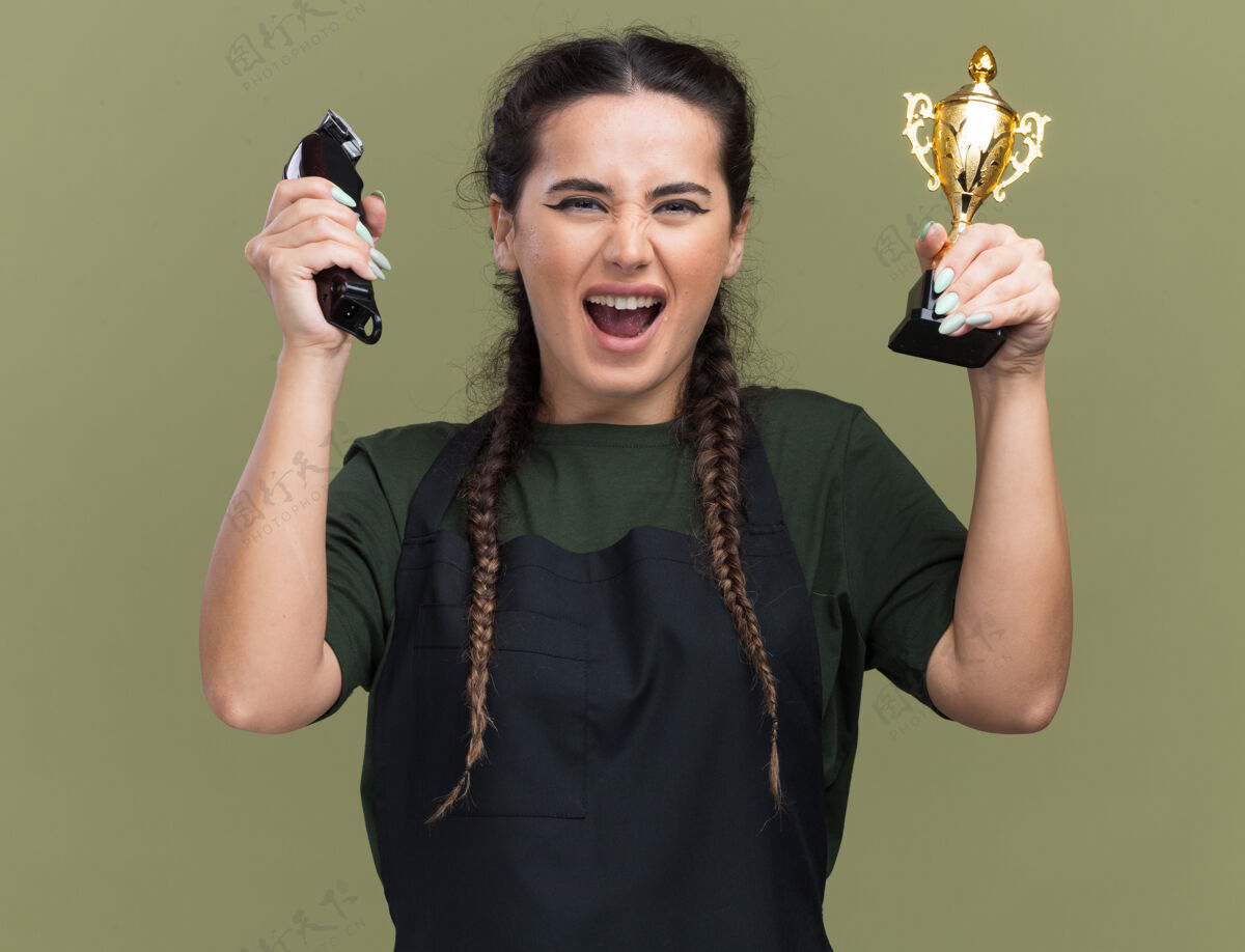 表情兴奋的年轻女理发师穿着制服拿着理发器和优胜者杯隔离在橄榄绿的墙上女性市民理发师