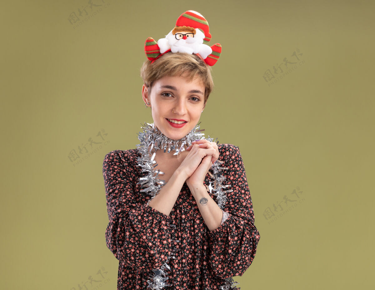 圣诞老人可爱的年轻女孩戴着圣诞老人的头带 脖子上戴着金属丝的花环 双手在橄榄绿的墙上相互隔离 留着复印空间保持年轻手