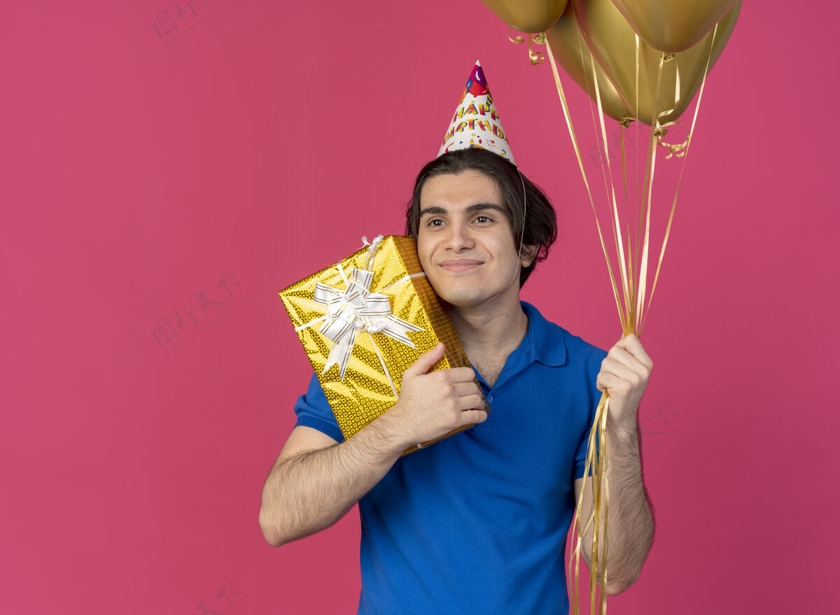 男人帅气的白种人戴着生日帽 手里拿着氦气球和礼盒看着身边盒子氦气粉色