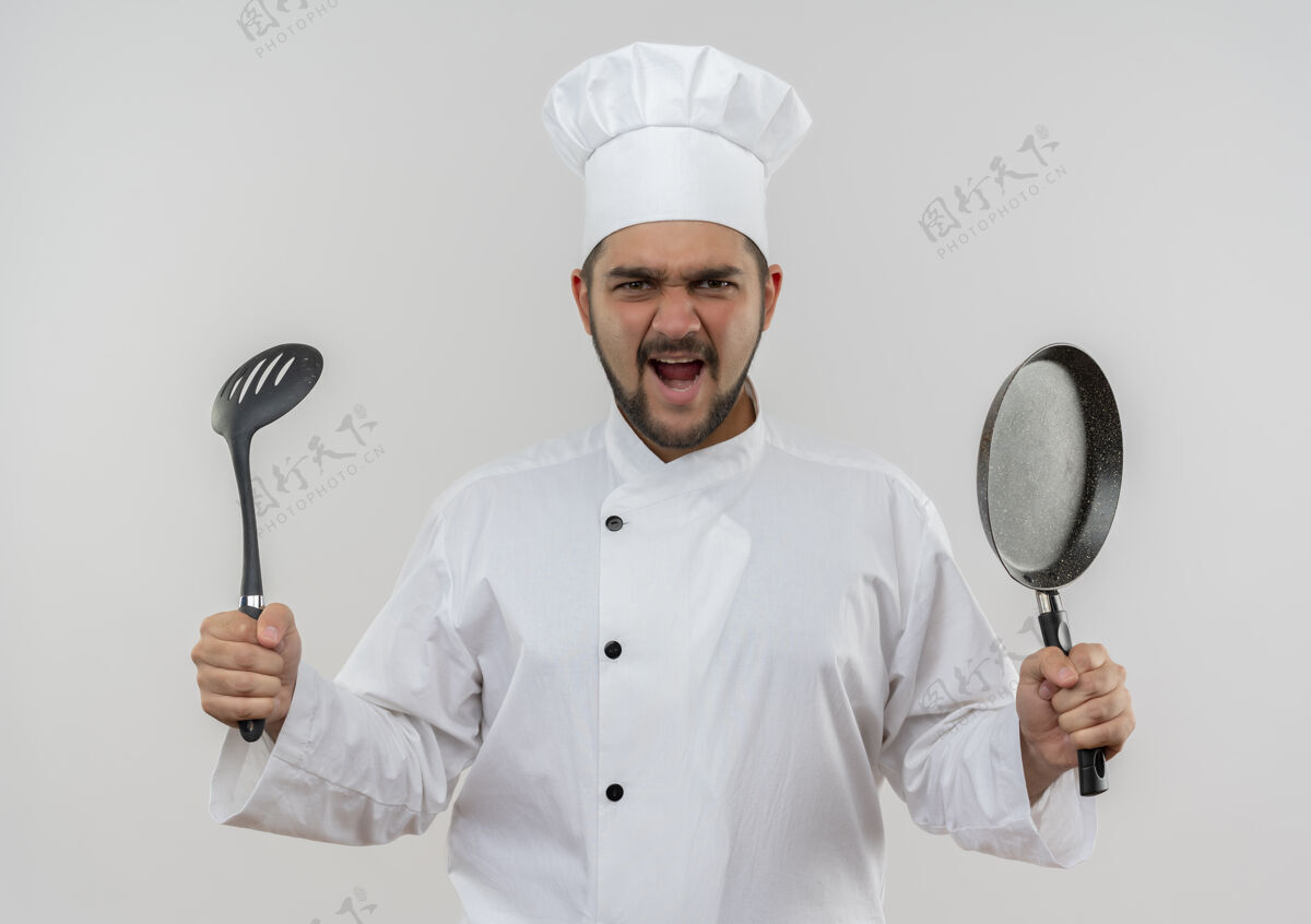 男性愤怒的年轻男厨师穿着厨师制服 拿着开槽汤匙和煎锅 隔离在白墙上年轻薯条愤怒