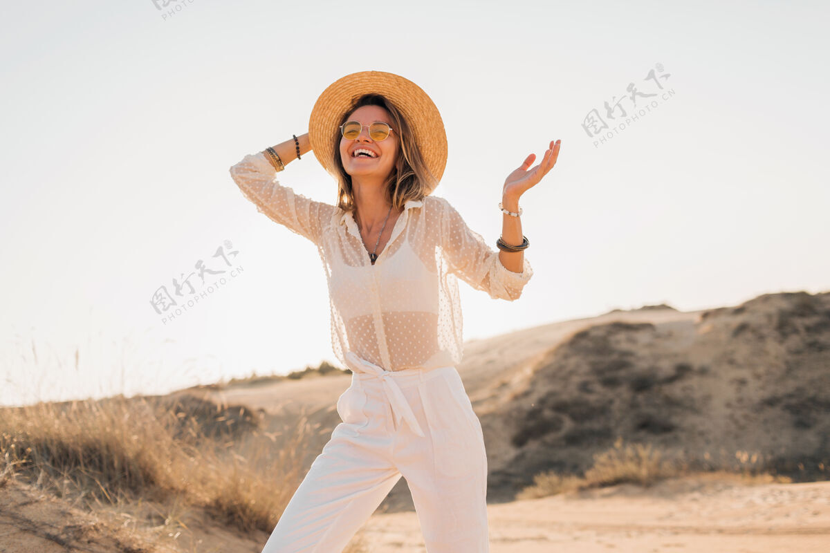 旅行时尚快乐的美丽微笑的女人在沙漠里摆姿势 穿着白色的衣服 戴着草帽和太阳镜在夕阳下太阳镜帽子时尚