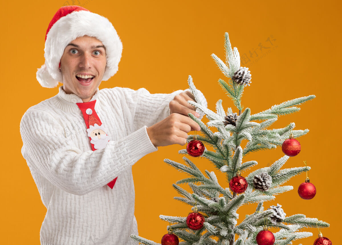 印象令人印象深刻的年轻帅哥戴着圣诞帽 打着圣诞老人的领带站在圣诞树旁 用圣诞球装饰圣诞树 看着隔离在橙色背景上的相机橙色装饰品圣诞老人