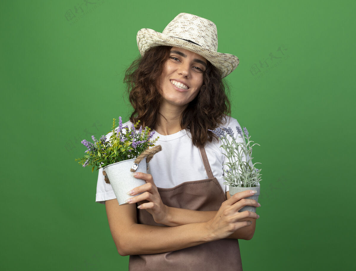 交叉微笑着的年轻女园丁 穿着制服 戴着园艺帽 手拿鲜花 在绿色的花盆里交叉花穿制服