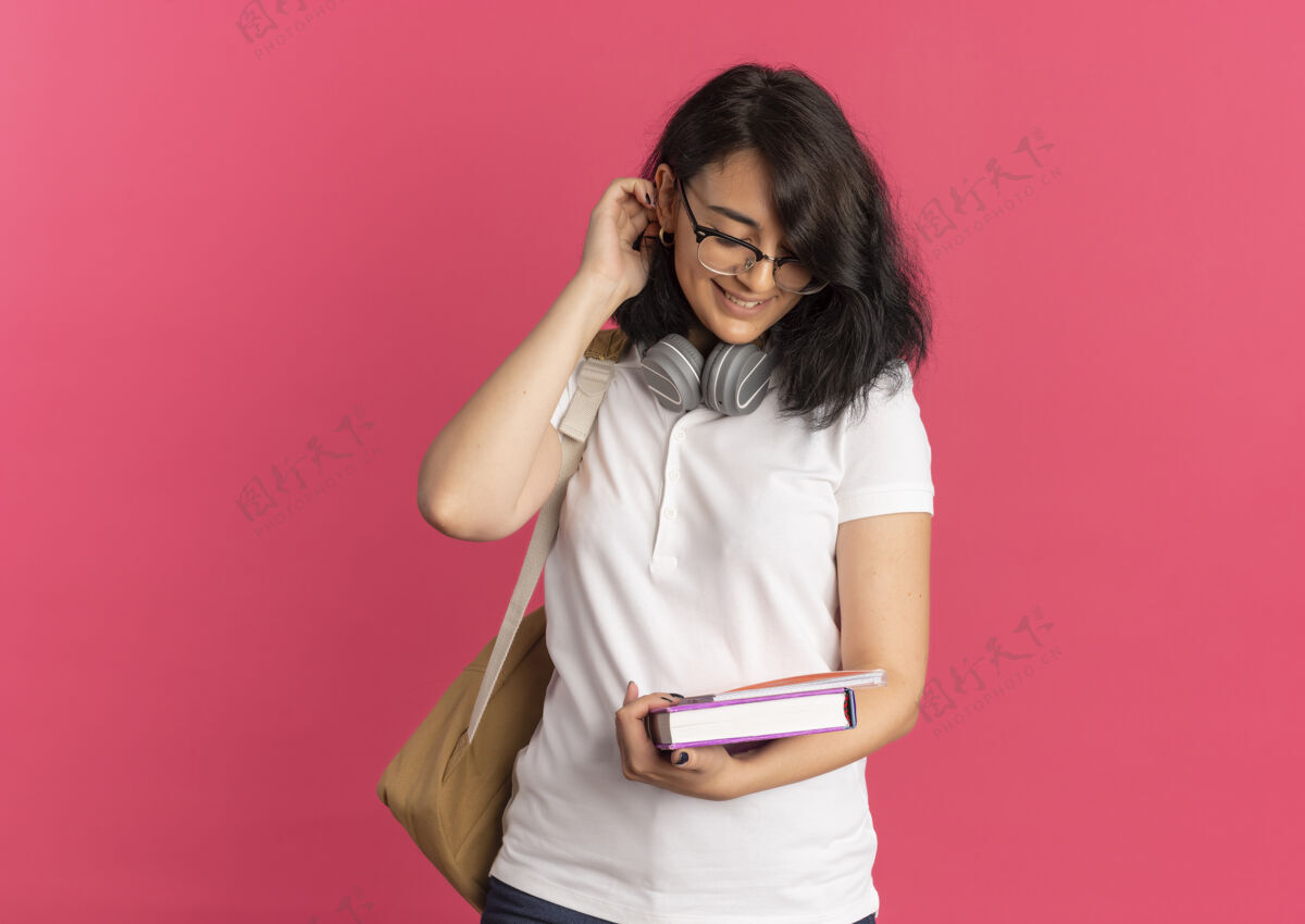 年轻年轻漂亮的高加索女生戴着眼镜 背着书包 戴着耳机 拿着书和笔记本看着粉红色的笔记本和复印空间穿复制包