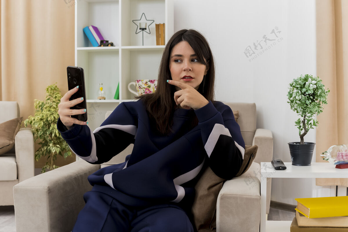 手机体贴的年轻漂亮的白人妇女坐在设计客厅的扶手椅上 拿着手机 看着手机 指着它漂亮住着扶手椅