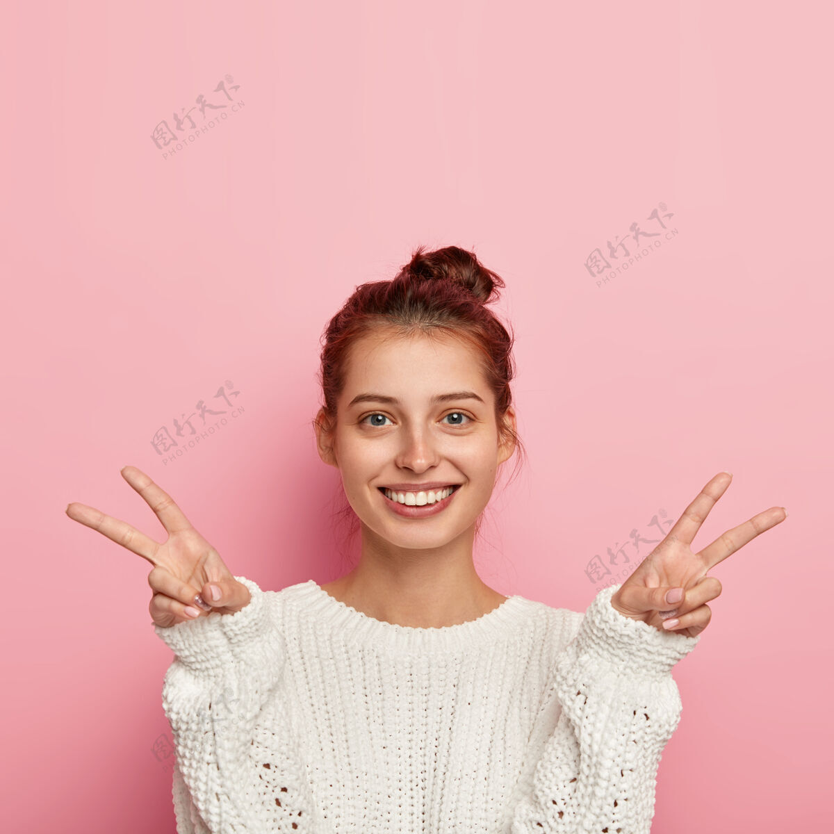 二喜洋洋的欧洲女人照片笑容灿烂 双手做出和平胜利的手势 有着温柔的神情 穿着白色针织毛衣 表达热情 显出幸福的迹象 孤立在粉色上女孩脸表情