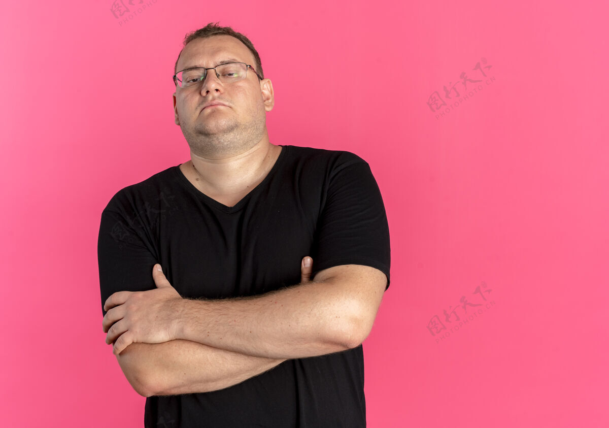 男人戴眼镜的超重男子身穿黑色t恤 面色凝重 双臂交叉放在胸前 站在粉红色的墙上站穿眼镜