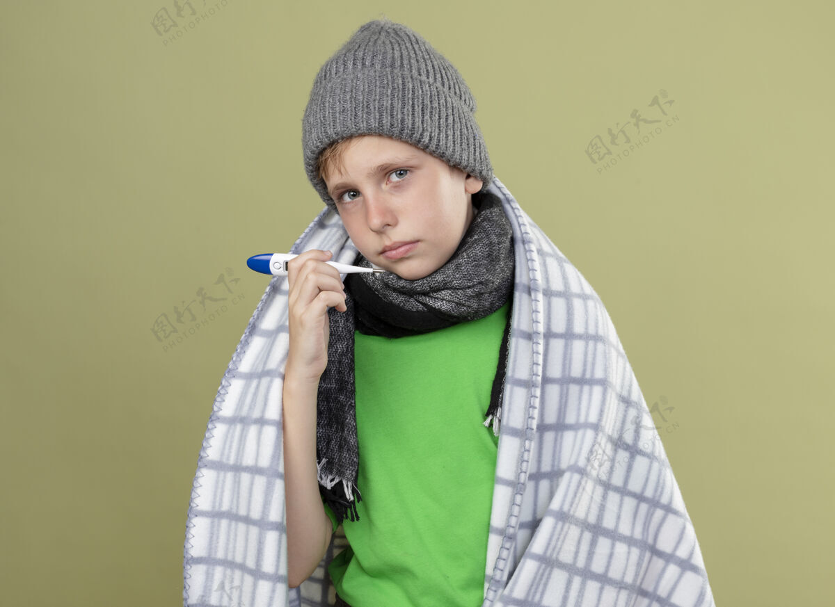 不开心生病的小男孩穿着绿色t恤 戴着暖和的围巾 帽子裹着毯子 站在光墙上 显示体温计不高兴和生病生病小灯