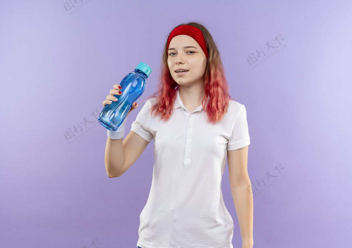 自信站在紫色的墙上 一个年轻的运动型女人拿着一瓶水 带着自信的微笑女人运动员运动