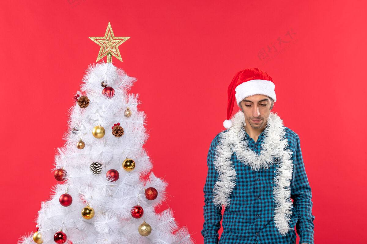 站着一个戴着圣诞老人帽子 穿着蓝色条纹衬衫 站在红色圣诞树旁低头的悲伤的年轻人礼物衬衫年轻