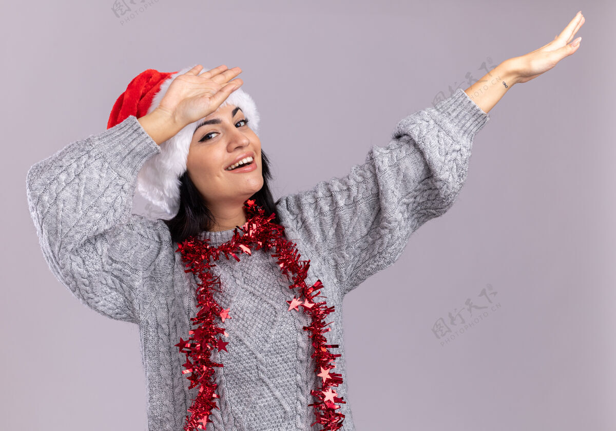 快乐快乐的年轻白人女孩戴着圣诞帽 脖子上戴着金箔花环 在白墙上做着一个孤立的手势金属丝圣诞节花环
