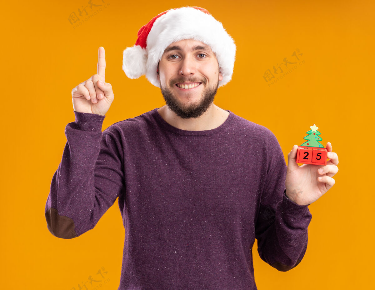 手指穿着紫色毛衣 戴着圣诞帽的快乐年轻人展示着玩具方块 25号微笑着 食指站在橙色的墙上圣诞老人站立二十