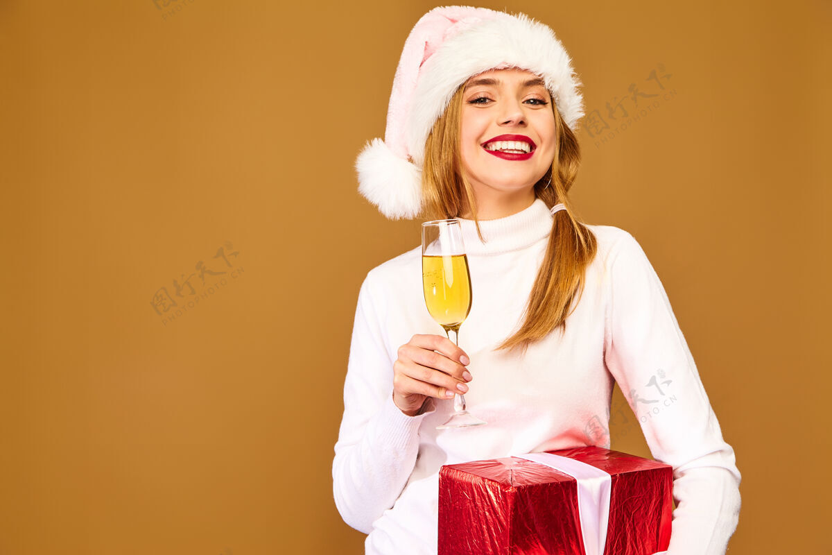 享受戴着圣诞帽和大礼盒的模特在金墙上喝香槟肖像人们圣诞