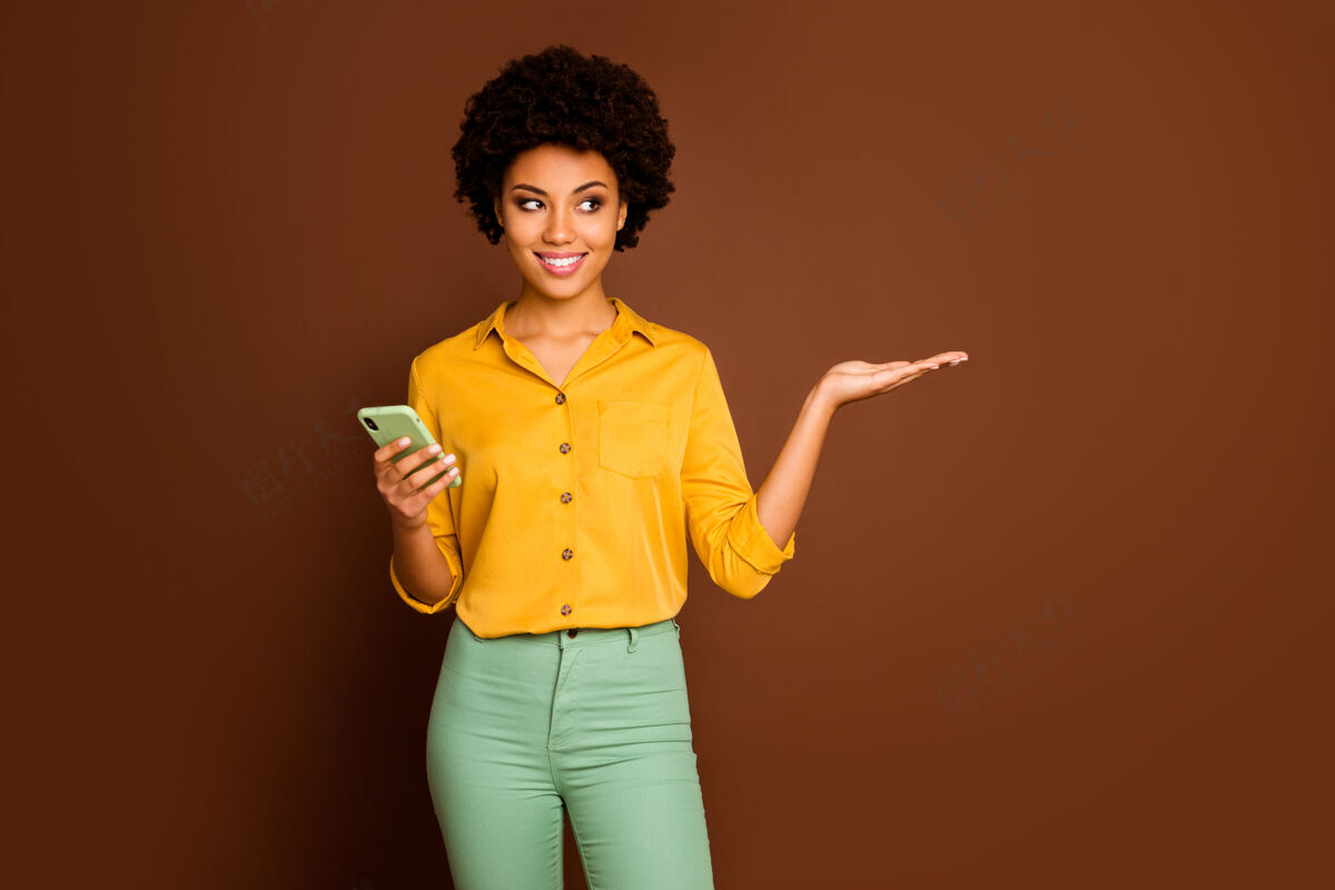 头发正面开朗的黑人女孩肖像使用智能手机牵手展示在线社交网络折扣推荐促销广告穿黄色套装孤立棕色微笑多国在线