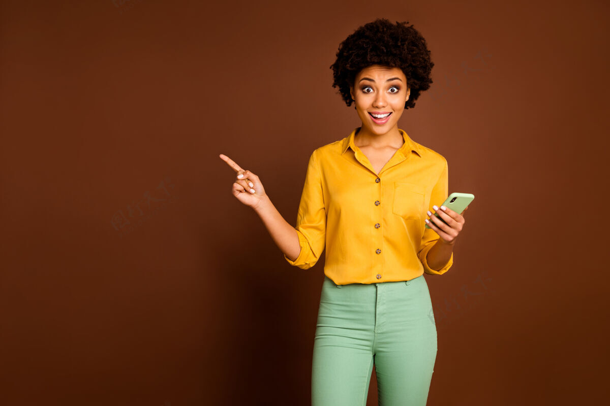 手机震惊疯狂的非裔美国女生用智能手机点指复制空间表明社交媒体销售尖叫哇穿黄绿色时髦裤子裤子孤立棕色信息惊喜手势