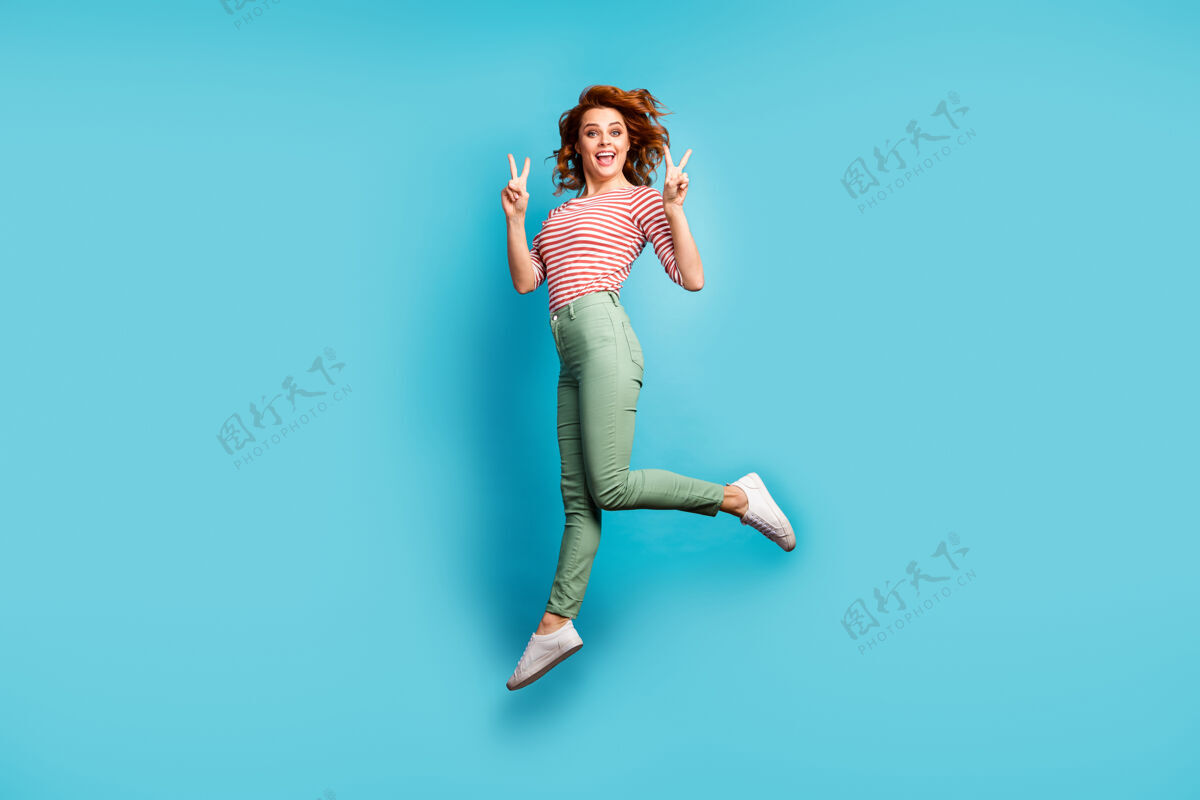 欢乐美丽的疯女人跳高的全身轮廓照片显示v形标志符号欢喜穿休闲红白衬衫绿裤子运动鞋孤立的蓝色乐趣女孩年轻