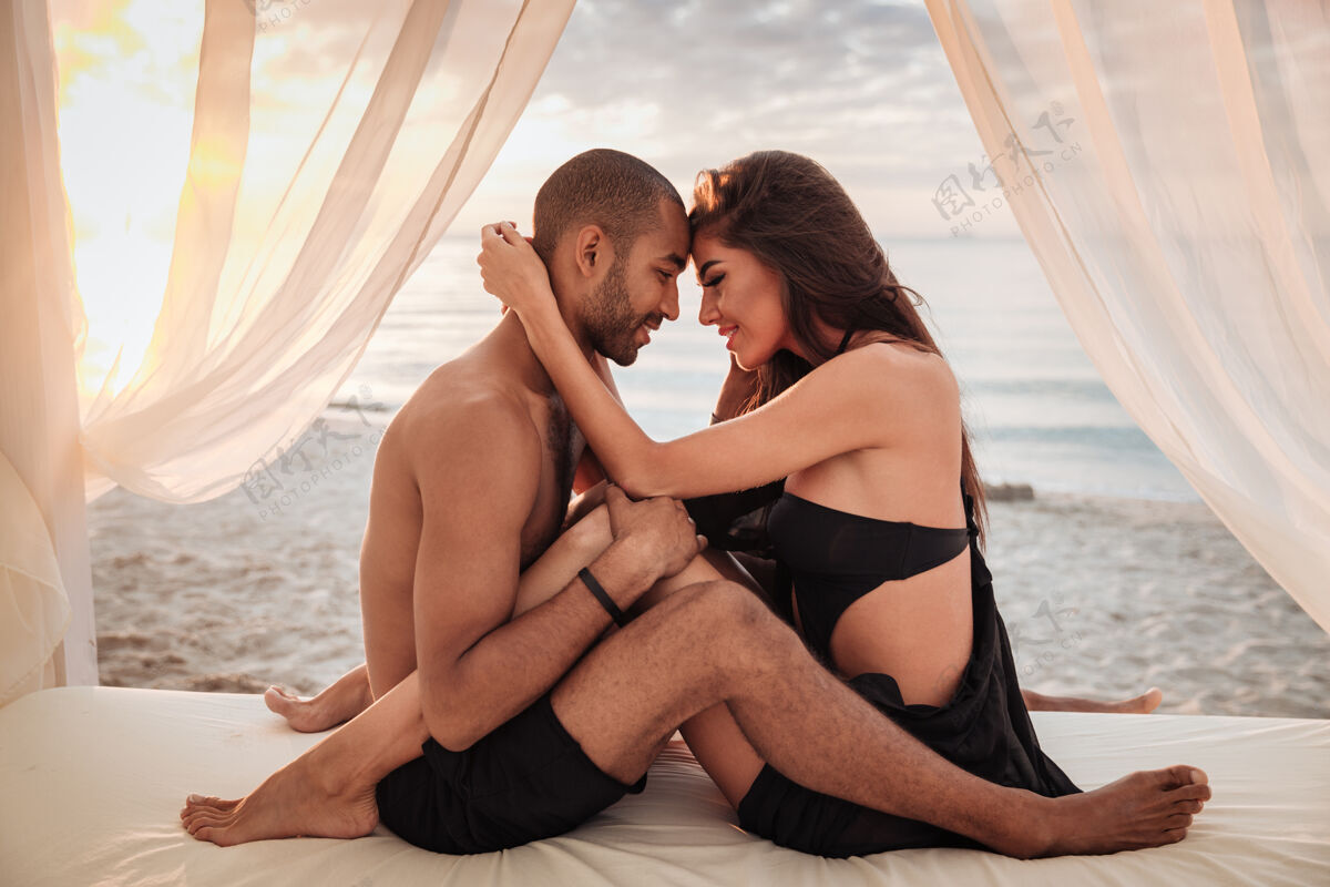 赤脚快乐的年轻夫妇坐在床上拥抱在沙滩上休闲情侣快乐