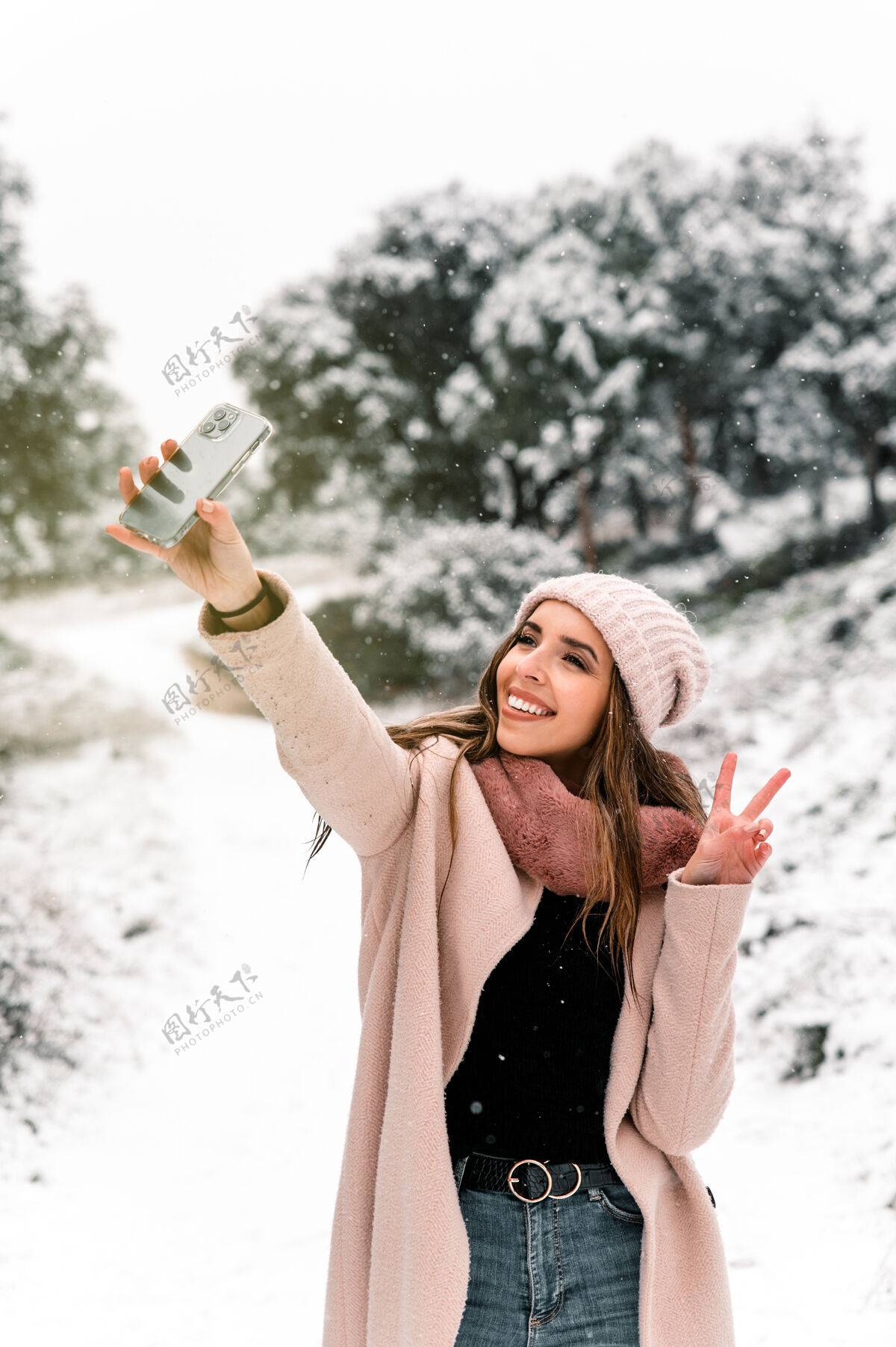 阴天穿着暖和外套的女人站在树林里 一边用智能手机自拍雪景漫步风景时尚