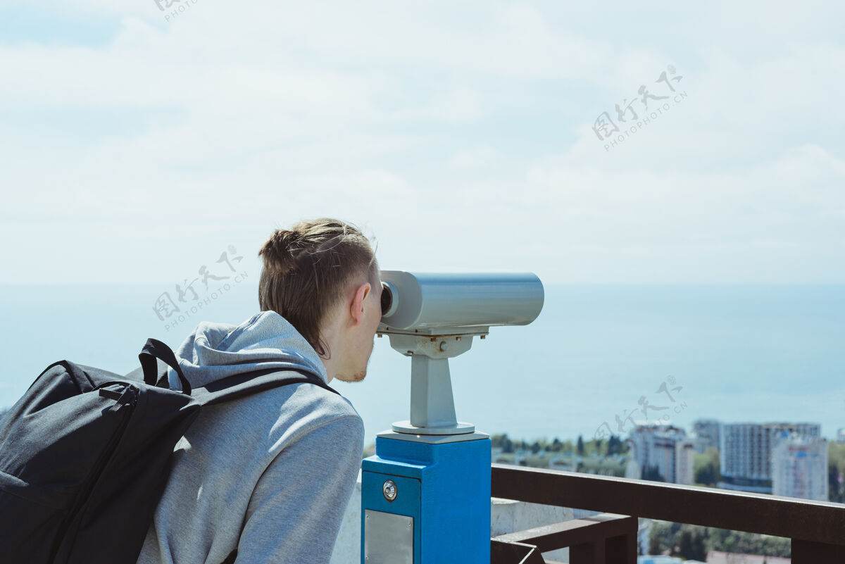 操作年轻的潮人旅游者透过金属硬币操作的双筒望远镜观看海 天和城市 横向生活方式股票图片游客冒险观光