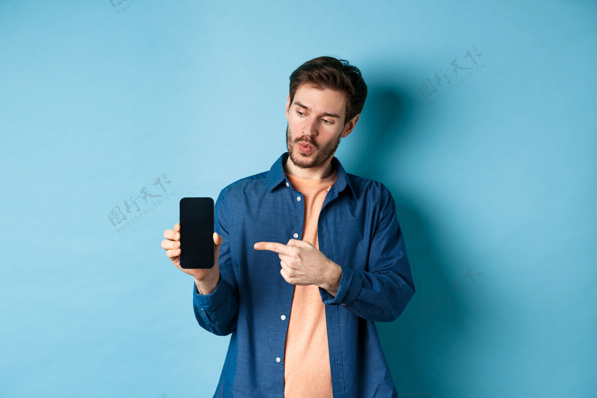 年轻帅哥谈论应用程序的画面 手指着手机屏幕 站在蓝色背景上屏幕酷胡须