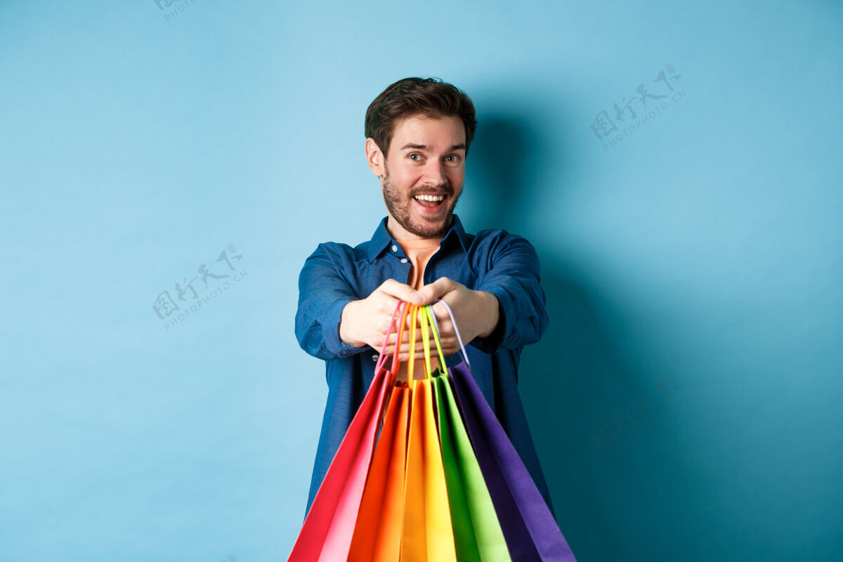 朋友开朗的小伙子手拿购物袋 面带微笑 站在蓝色的背景上给你送礼物表情肖像购物者