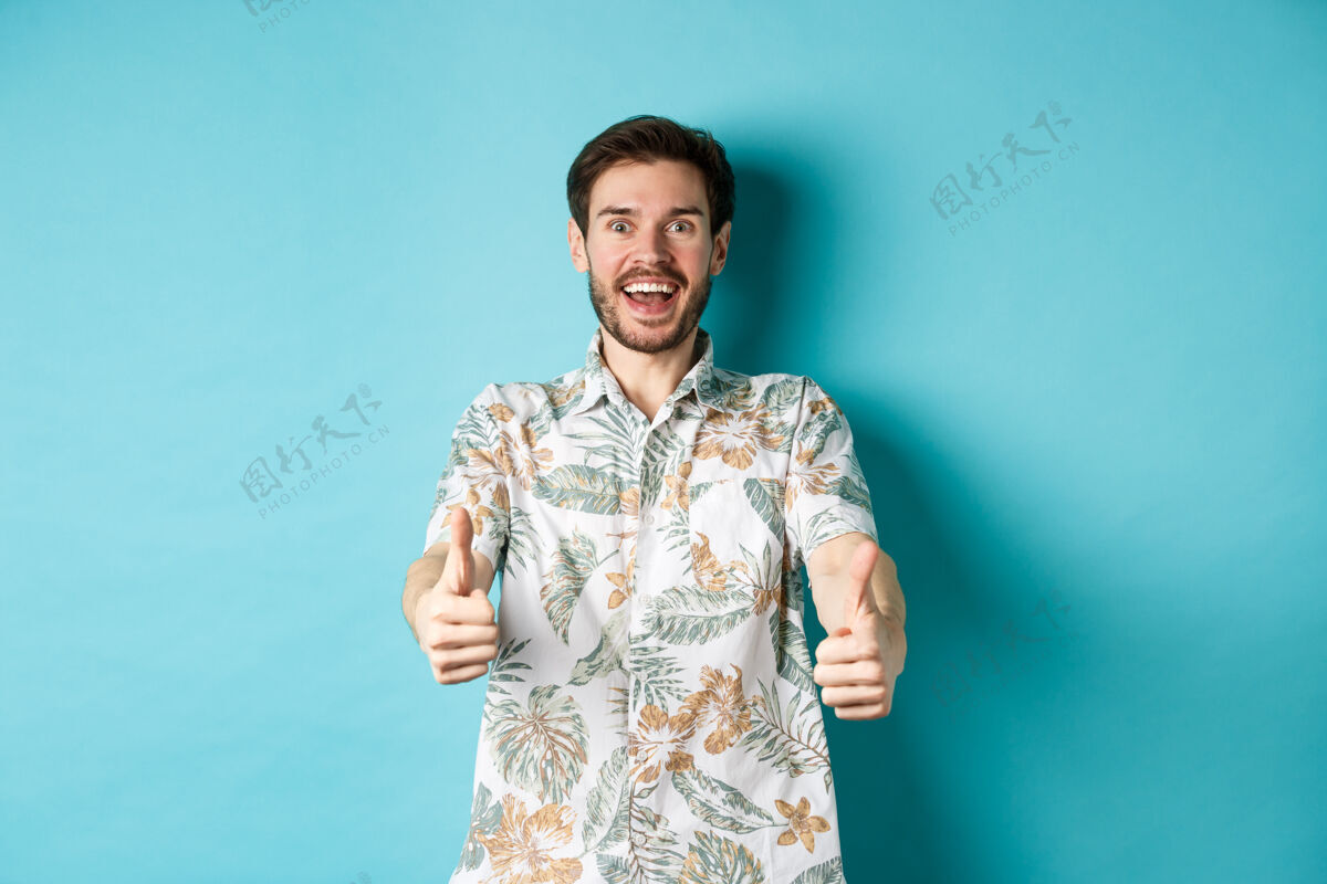 休闲穿着夏威夷衬衫的兴奋的微笑着的家伙竖起大拇指 看起来很惊讶 看看酷的宣传片 站在蓝色的背景上男人酷成人