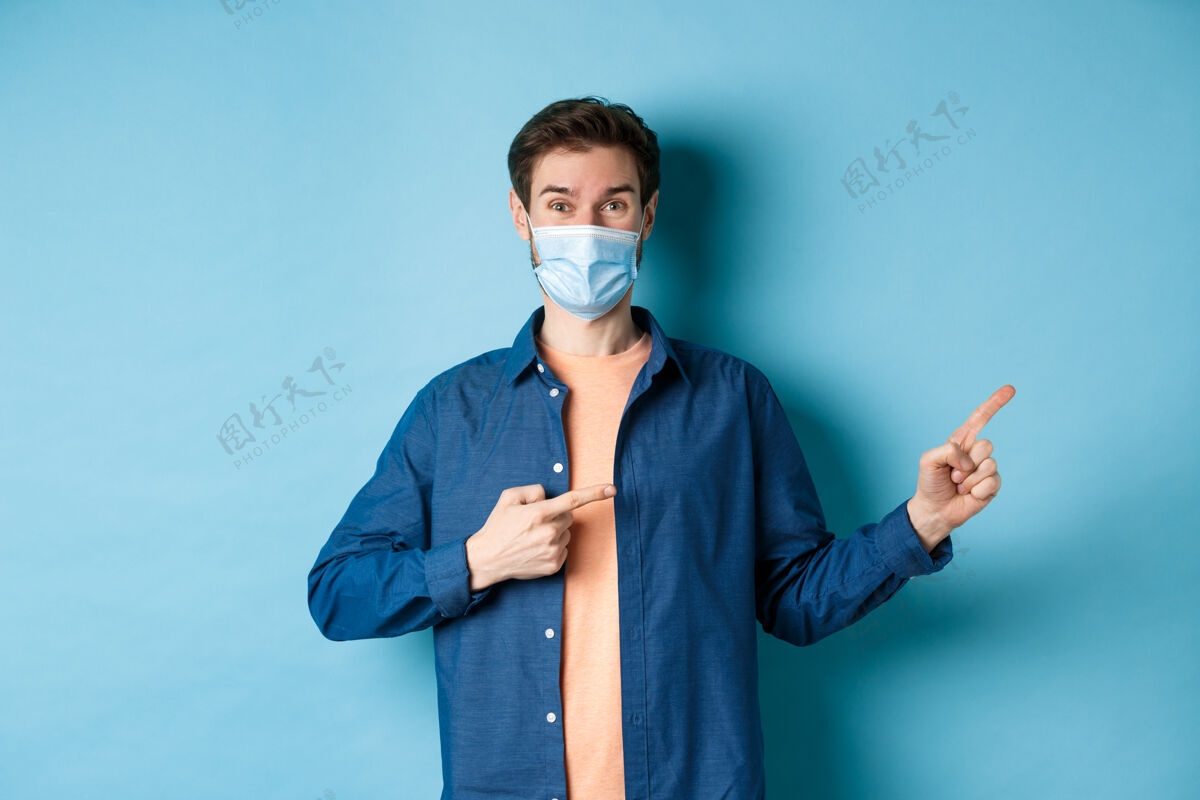 冠状病毒Covid-19与大流行概念戴着医用口罩的快乐健康的家伙手指着右上角 展示着广告 站在蓝色的背景上情感男人成人