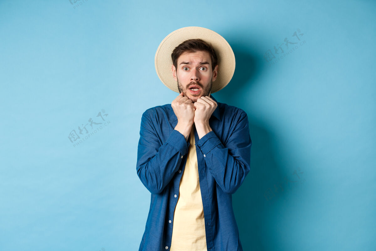 年轻年轻的游客吓得浑身发抖 看着暑假里可怕的东西 戴着草帽 站在蓝色的背景上自信人帽子