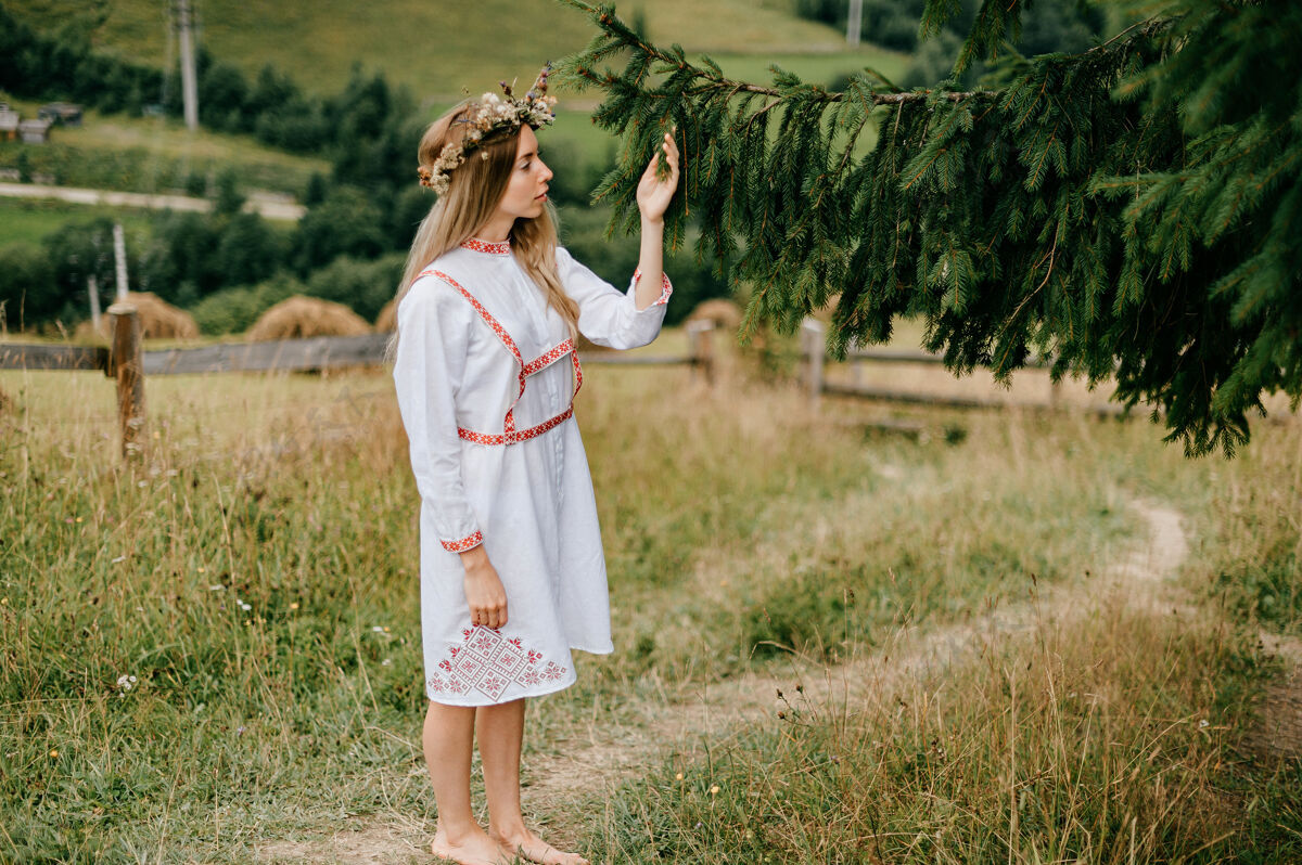 风景年轻迷人的金发女孩 身穿白色连衣裙 头上戴着刺绣和花环 在乡村风景中与冷杉枝合影花环女孩金发