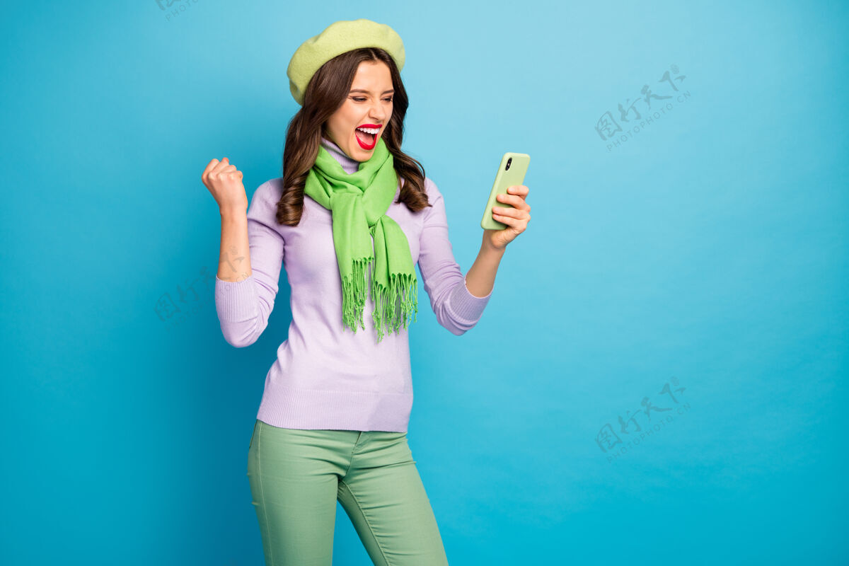 时尚美女手握电话看新帖正面评论反应穿绿色贝雷帽紫色高领围巾裤子隔离蓝色墙壁网络手机法国