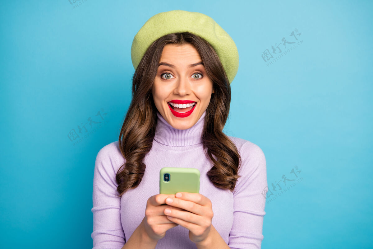 互联网惊喜积极时髦的女孩使用智能手机阅读社交媒体新闻印象深刻尖叫哇哦omg穿少女风格时尚紫色套装头饰隔离蓝色墙壁电话信息欢乐