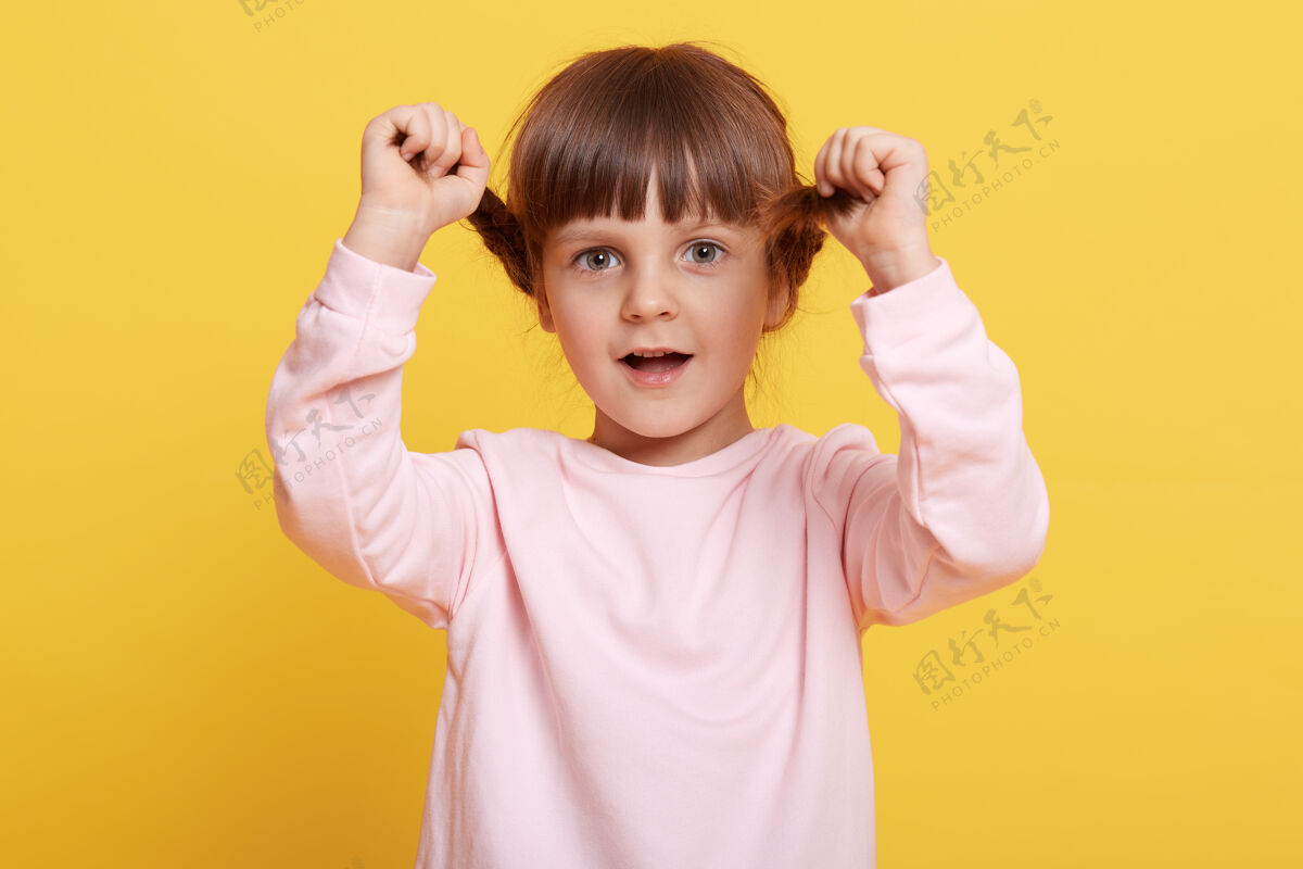 毛衣快乐的小女孩扎着辫子 张着嘴直视镜头 手放在辫子上 穿着淡粉色的休闲装 在黄色背景下摆出孤立的姿势活动情感乐观