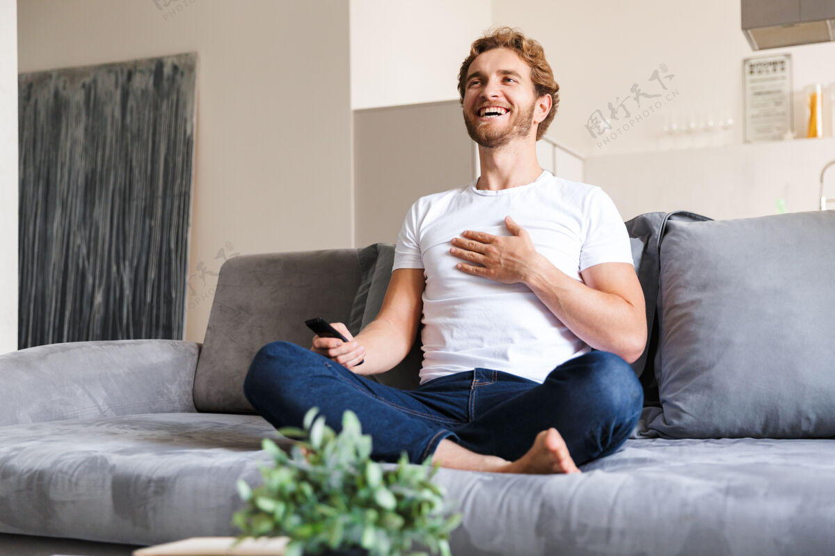 国内一个英俊快乐的年轻人在家里的沙发上拿着遥控器看电视舒适休闲男人