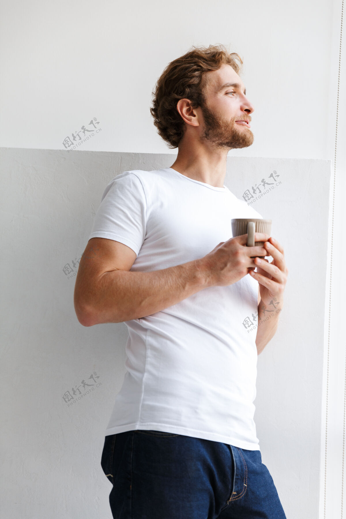 肖像微笑的年轻人端着一杯茶 站在家里的窗前享受男人马克杯