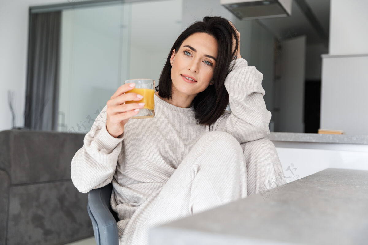 饮料高兴的女人30多岁喝橙汁 而在明亮的现代房间休息深褐色成人沙发