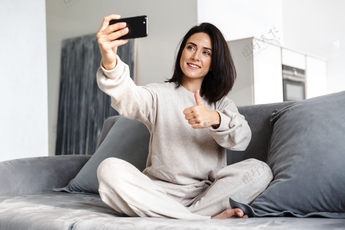 女人30多岁的傻女人坐在明亮公寓的沙发上用手机自拍舒适室内成熟