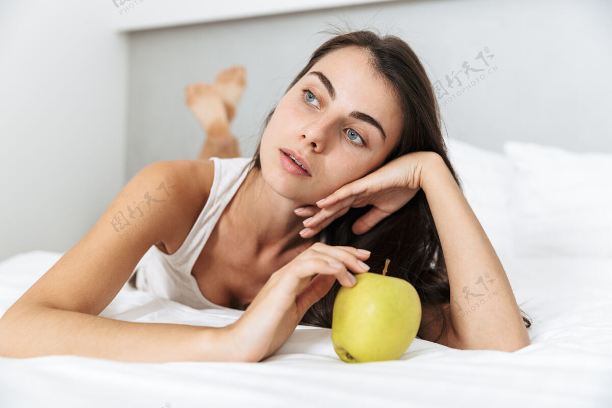 休闲美丽的年轻女子在家里的床上放松 手里拿着一个青苹果快乐女人成人