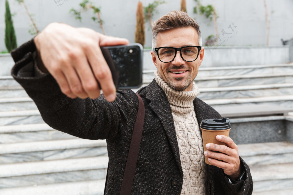 享受穿着夹克的帅哥坐在户外自拍 手里拿着一杯外卖咖啡肖像设备吸引