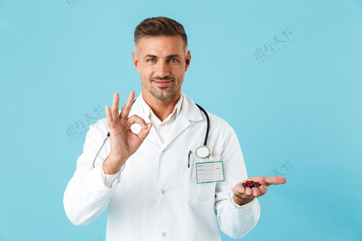 中年人一个英俊的成熟男子医生在蓝色的墙壁上摆姿势 拿着药片和维生素 显示出良好的姿态表情药物工人