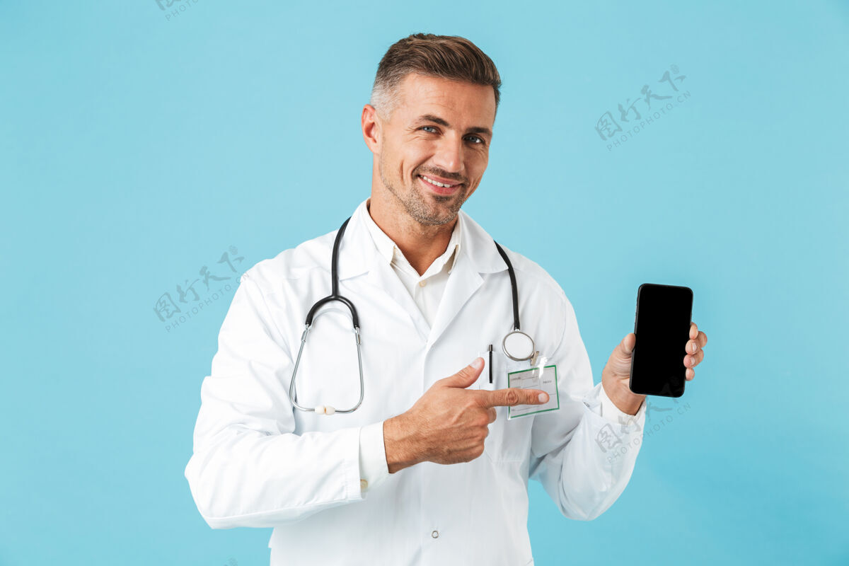 治疗微笑的医生身穿白大褂 手持听诊器手持手机 孤立地站在蓝色的墙上工人听诊器心脏病专家