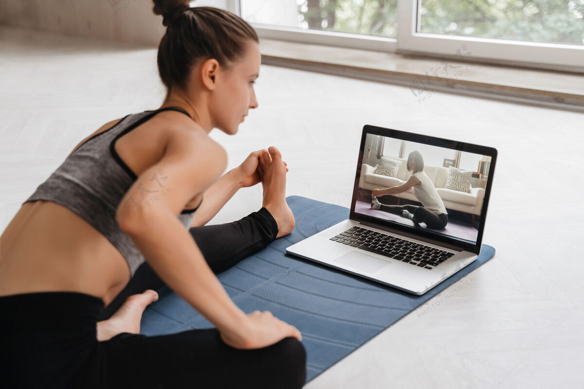 远程适合穿着运动服的女士在家里用笔记本电脑在运动垫上做瑜伽教程.女教练在电脑上有一个虚拟瑜伽课女人体操私人教练