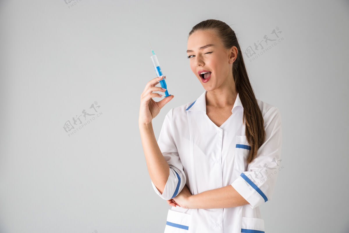 嘴唇照片中 一位年轻漂亮的美容师医生隔着灰墙拿着注射器眨眼女人专家制服