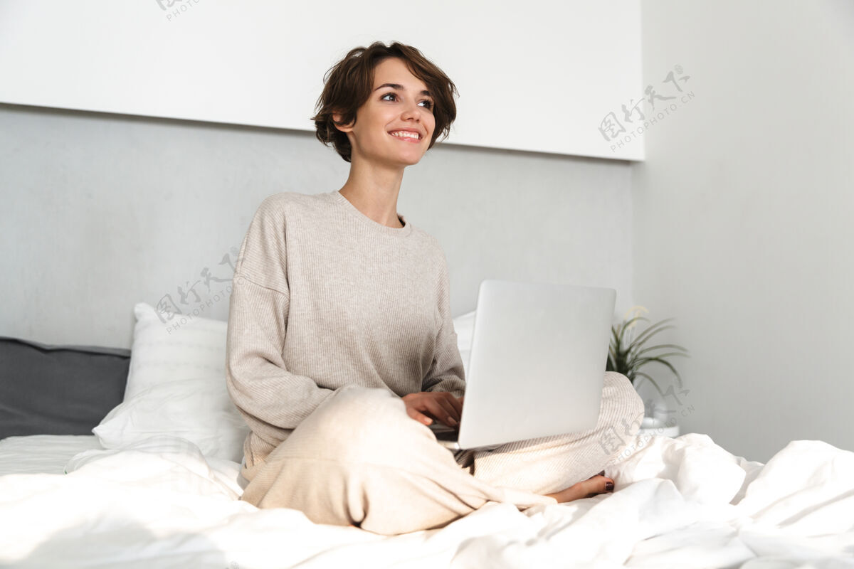 宁静微笑的年轻女孩 早上在床上放松 用笔记本电脑打字枕头室内
