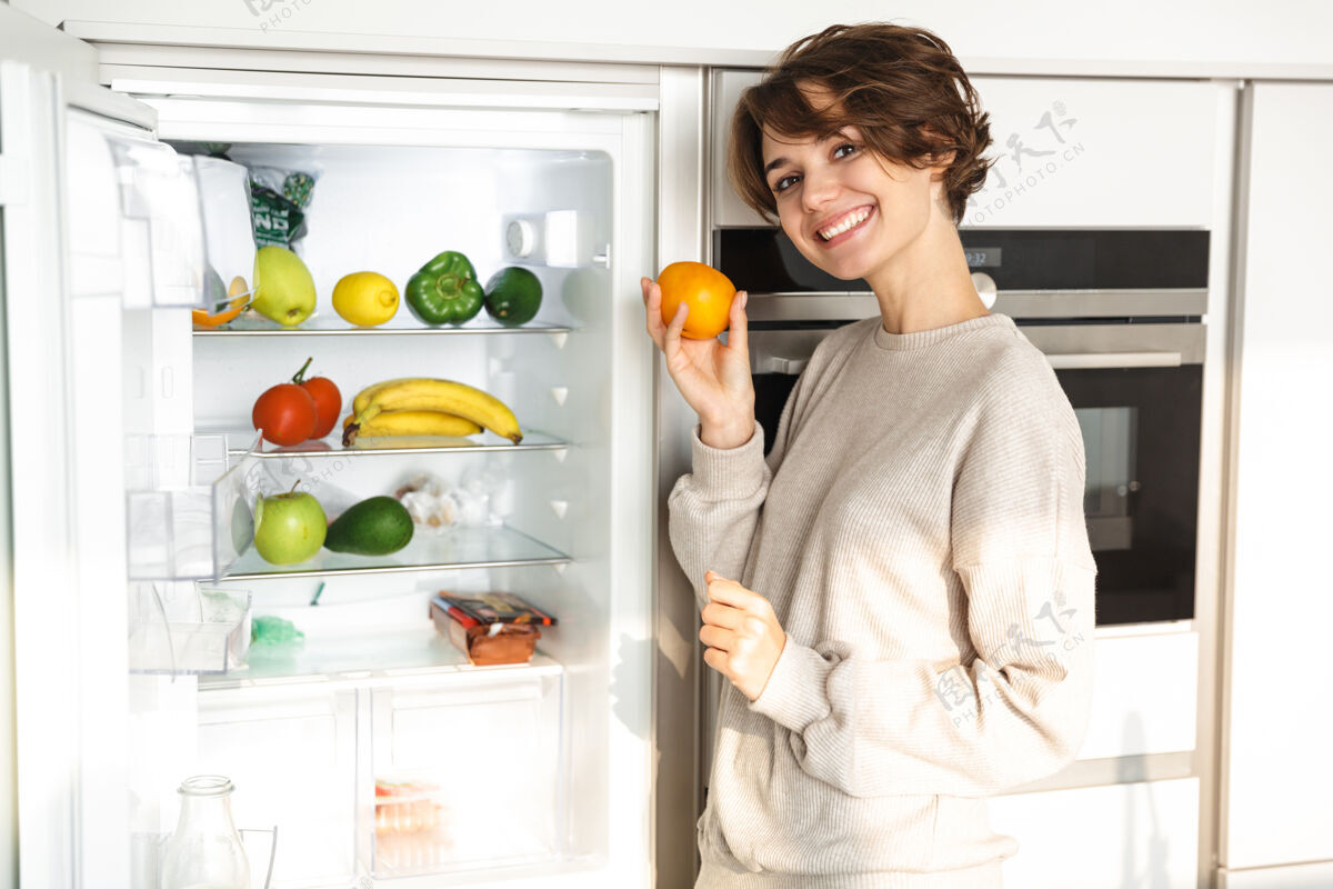 成人微笑的年轻女子站在冰箱前 手里拿着橘子冰箱蔬菜漂亮