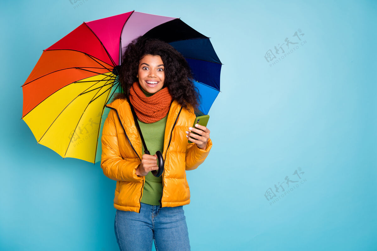 多民族深色皮肤的女士带着雨伞在蓝色背景的智能手机上浏览互联网短信年轻微笑