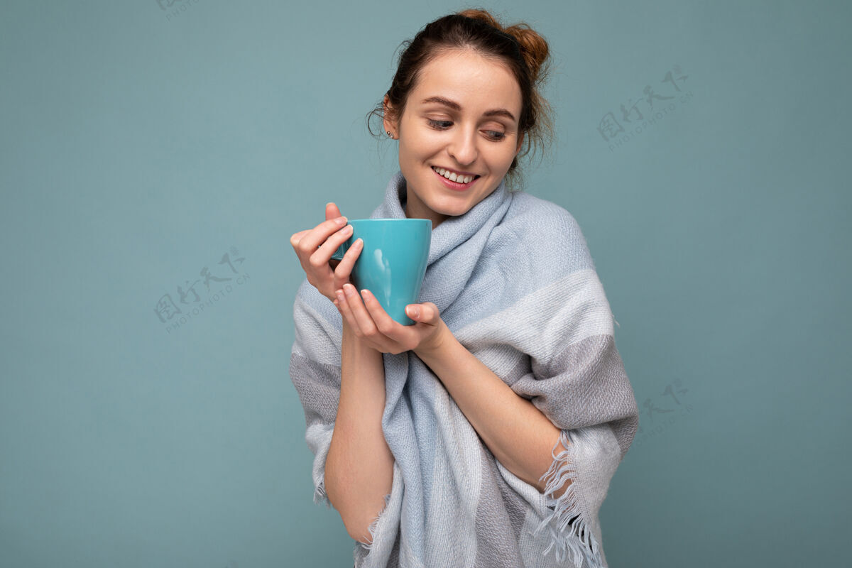茶这张照片拍摄的是一位年轻可爱的黑发女子 她戴着温暖的蓝色围巾 在蓝色的背景下 拿着蓝色的马克杯 喝着咖啡 低头看美味的口渴冰沙