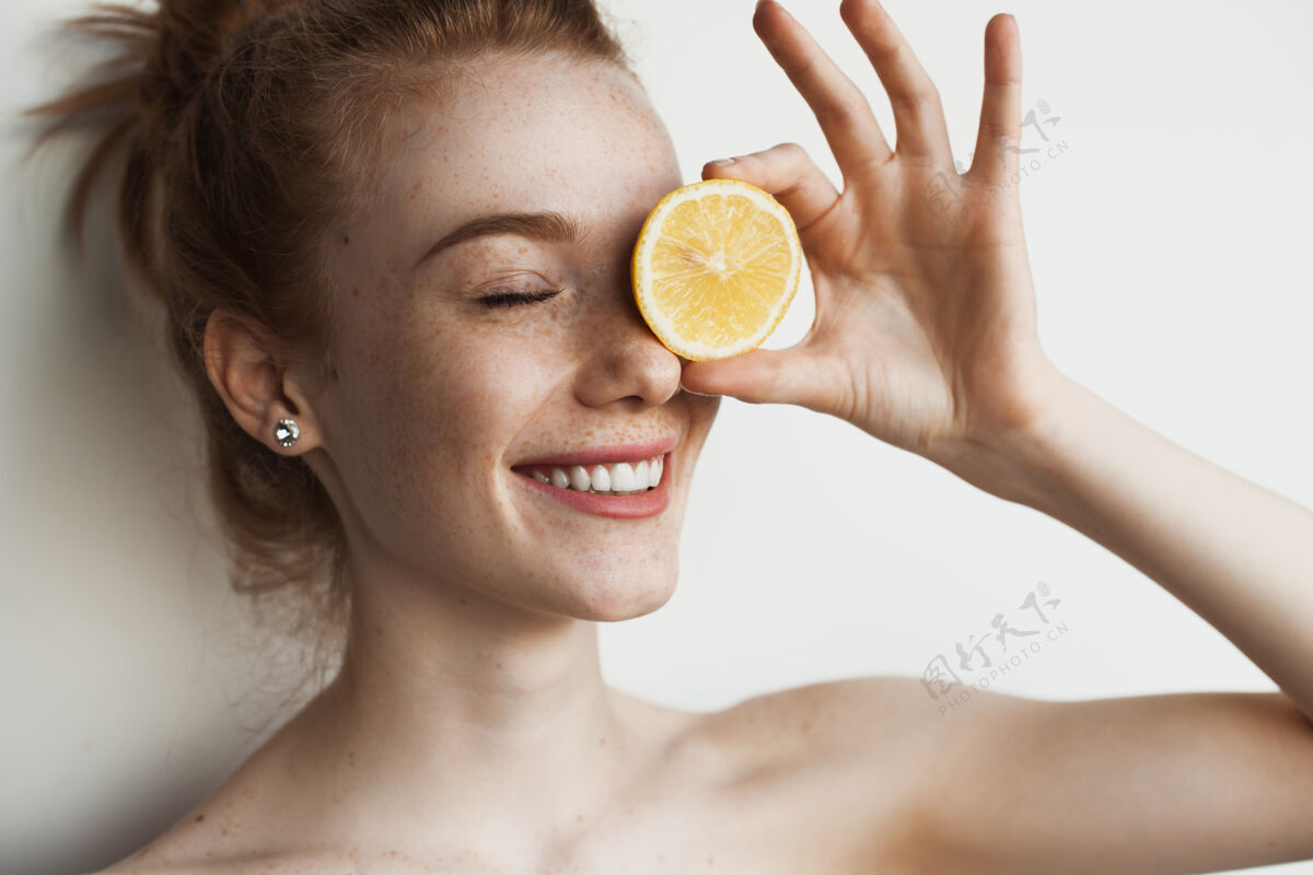 健身一个长着雀斑的姜黄色高加索女人用柠檬遮住眼睛 在白色的画室墙上微笑着夏天年轻创意