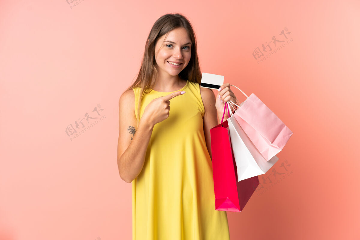 付款一位年轻的立陶宛妇女 手拿购物袋和信用卡 被隔离在粉色背景下购买年轻银行